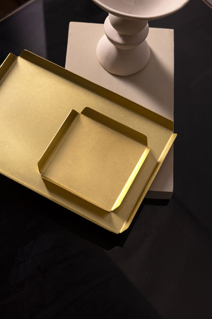 Pictured is a square 100mm X 100mm real brass tray with raised, bevelled edges to each side. The tray is sitting upon another, medium sized brass tray in the same design with bevelled edges but larger. The trays are placed landscape across the white block, with the base of a white, thin vase barely in frame toward the top of the picture. The white vase, white block and brass trays are upon a black, reflective table top. The brass tray is brushed and matte and is made by Empire Copper.